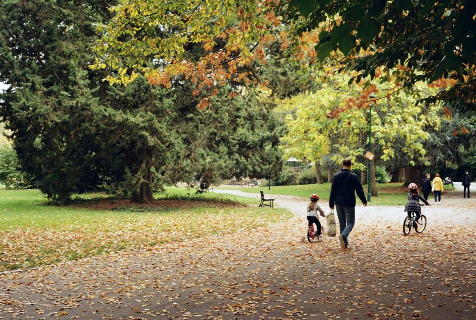 A person, possibly a parent, walking with two young children on a paved pathway within Battersea Park during autumn. The person is carrying a white bag and is dressed in dark clothing, while one child