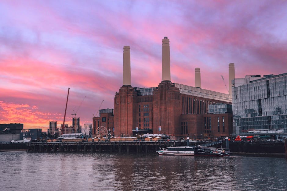 A low-angle view of Battersea Power Station's exterior during daytime, featuring a large brick facade with vertical windows and shadowed areas, alongside a tall, cylindrical chimney rising into a clea